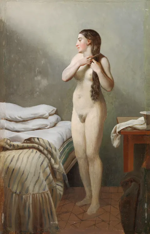 A female standing by a bed, combing her hair.